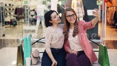 漂亮的年轻女士正在一起自拍，使用智能手机坐在购物中心休息和玩得开心
